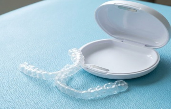 A set of transparent, invisible orthodontic aligners resting next to their white protective case on a light blue surface.